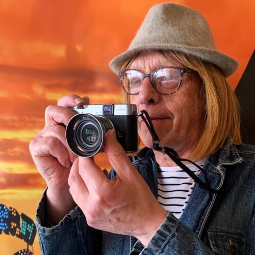 Kate, wearing a trilby hat and holding her camera mid photo taking